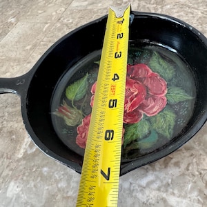 Könnte beinhalten: Schwarze gusseiserne Bratpfanne mit einem bemalten Blumendesign mit roten Rosen und grünen Blättern. Die Bratpfanne hat einen Durchmesser von ca. 15 cm, gemessen mit einem gelben Maßband.