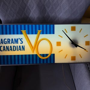 Puede incluir: Reloj rectangular iluminado con el texto "Seagram's Canadian" y un logotipo dorado "V0". El lado izquierdo tiene rayas verticales azules. La esfera del reloj es blanca con marcadores de hora cuadrados naranjas y manecillas negras.