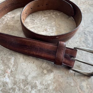 May include: A brown leather belt with a silver-colored buckle. The belt has a worn appearance, with visible creases and variations in color. The buckle is rectangular with a rounded edge.