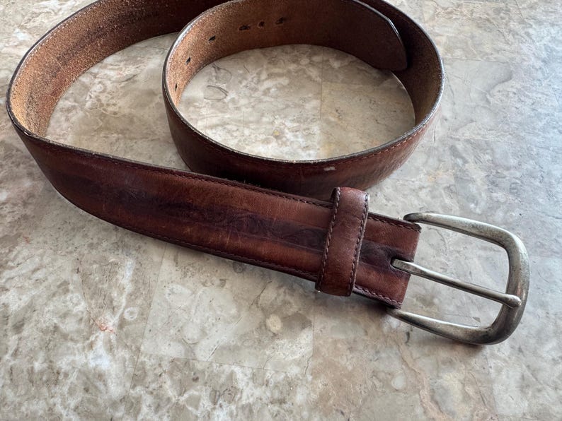 May include: A brown leather belt with a silver-colored buckle. The belt has a worn appearance, with visible creases and variations in color. The buckle is rectangular with a rounded edge.