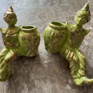 May include: A pair of lime green ceramic figurines, each featuring a seated figure attached to a hollow vessel. The figures have pointed headpieces and are decorated with gold-toned accents on their clothing and the textured surfaces of the pots.
