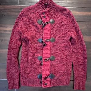 May include: A burgundy knit sweater with a high collar and toggle closures. The sweater has a textured appearance and features black toggle clasps down the front. The sleeves and shoulders have a slightly darker shade of burgundy.