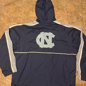 Op de afbeelding: Marineblauwe jas met capuchon en lichtblauwe en witte accenten. Voorzien van een groot lichtblauw "NC" logo op de rug. De jas heeft een trekkoord aan de onderkant en lichtblauwe strepen op de mouwen.