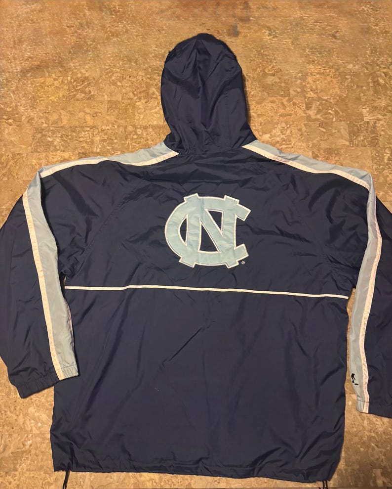 Op de afbeelding: Marineblauwe jas met capuchon en lichtblauwe en witte accenten. Voorzien van een groot lichtblauw "NC" logo op de rug. De jas heeft een trekkoord aan de onderkant en lichtblauwe strepen op de mouwen.