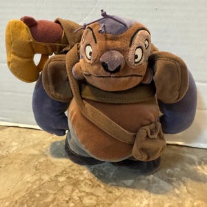 May include: A plush toy of a character with brown fur, purple accents, and large ears. The toy has a satchel and a large, yellow, claw-like appendage. The character has a grumpy expression and is standing on a patterned surface.
