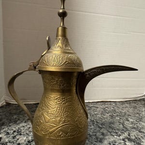 May include: A vintage brass coffee pot with detailed floral and geometric patterns. It has a long, curved spout, a handle, and a decorative lid with a finial. The brass has a warm, aged patina, indicating its use and age.