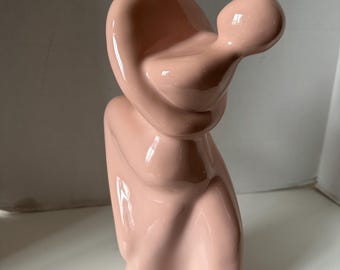 May include: A pink ceramic sculpture of two figures embracing. The figures are abstract and stylised, with smooth, rounded forms.