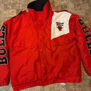 May include: Red Chicago Bulls jacket with black and white accents. The jacket features the team name "BULLS" in large black letters on the sleeves. The Chicago Bulls logo is on the chest. The jacket has a black collar and a button-up front.