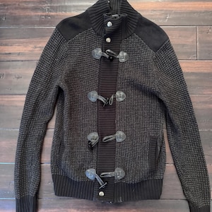 May include: A dark brown and black knit sweater with a high collar and toggle closures. The sweater features a textured pattern on the body and solid black accents on the shoulders, collar, and cuffs. The toggle closures are a dark brown color.