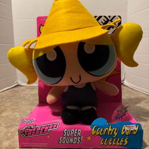 Puede incluir: Un peluche de Burbuja de Las Supernenas, con un sombrero amarillo y un atuendo negro. El juguete está en una caja rosa con el texto "SUPER SOUNDS! Country Band Bubbles". El juguete tiene grandes ojos azules y coletas amarillas.