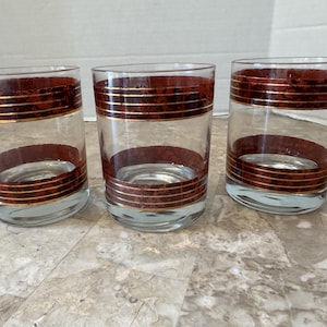 Può includere: Set di tre bicchieri da whisky in vetro trasparente con un motivo marrone e oro. I bicchieri hanno una base larga e un lato dritto.