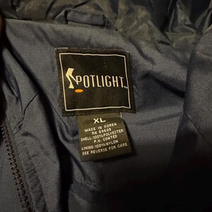 Op de afbeelding: Close-up van de binnenkant van een marineblauwe jas met een Spotlight-label. Het label bevat de tekst "Spotlight" en details over de materialen en herkomst van de jas, waaronder "Made in Korea" en de maat "XL".
