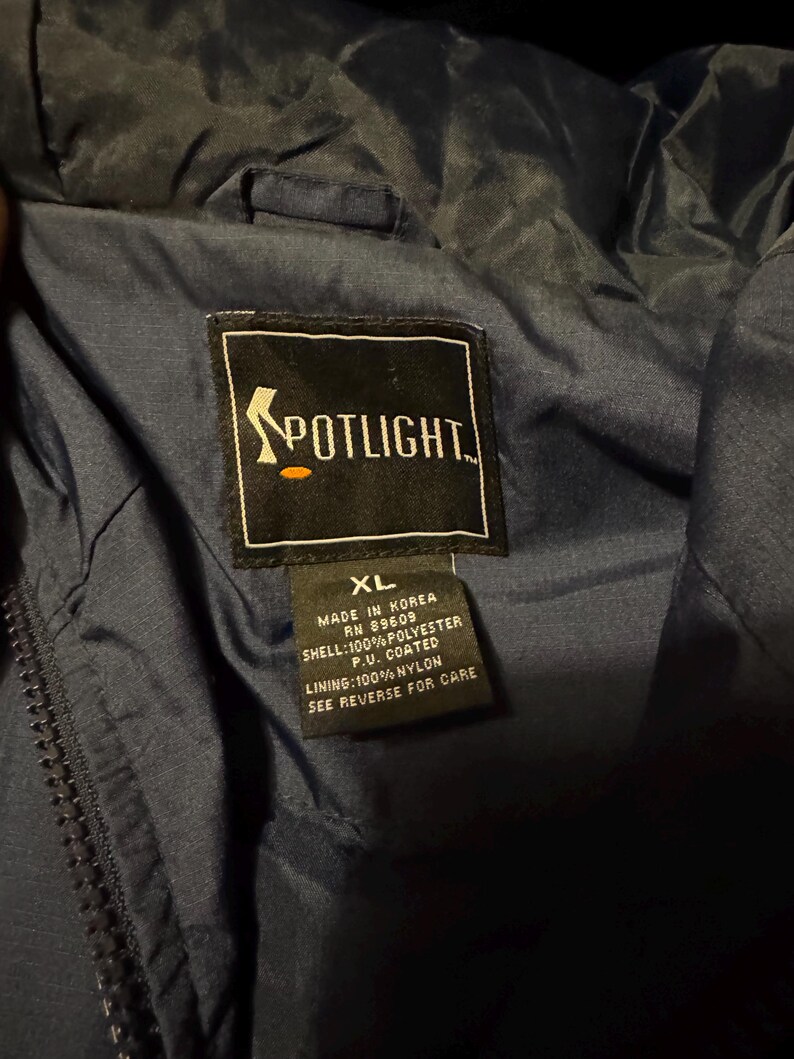 Op de afbeelding: Close-up van de binnenkant van een marineblauwe jas met een Spotlight-label. Het label bevat de tekst "Spotlight" en details over de materialen en herkomst van de jas, waaronder "Made in Korea" en de maat "XL".