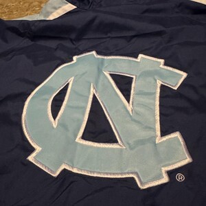 Op de afbeelding: Marineblauwe jas met een lichtblauwe en witte accentstreep. De jas heeft een groot lichtblauw geborduurd logo met een witte omtrek. De jas heeft een capuchon en een geregistreerd handelsmerk.