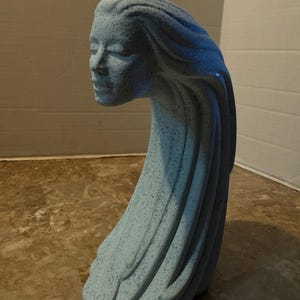 Puede incluir: Una escultura de cerámica azul claro que representa el rostro de una mujer y cabello fluido. La escultura tiene una textura moteada y una forma curva y alargada. El rostro de la mujer es sereno, con los ojos cerrados y una expresión suave. Es una pieza de arte decorativa.