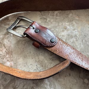 May include: A brown leather belt with a silver-colored buckle. The belt has a distressed appearance with visible wear and tear. The leather is a medium brown color with a slightly textured surface. The buckle is rectangular and has a classic design.