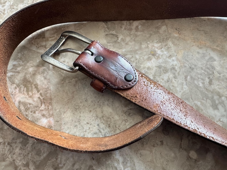 May include: A brown leather belt with a silver-colored buckle. The belt has a distressed appearance with visible wear and tear. The leather is a medium brown color with a slightly textured surface. The buckle is rectangular and has a classic design.