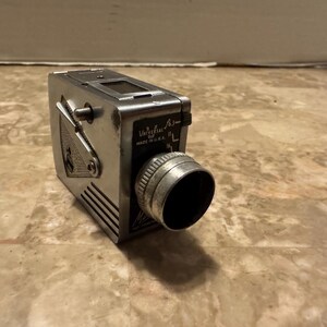 May include: Vintage Universal A-2 movie camera, made in the U.S.A. The camera is silver-toned with a lens, viewfinder, and various dials and levers. The camera has a rectangular body with a ribbed side panel.