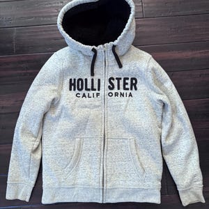 May include: A light gray zip-up hoodie with a black interior lining and hood. The hoodie features the text "HOLLISTER CALIFORNIA" in black block letters across the chest. It has two front pockets and a drawstring.
