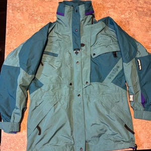 May include: A teal and blue vintage jacket with a high collar and multiple pockets. The jacket features the word "equipe" on the sleeve and purple accents on the collar and cuffs. The jacket is made of a lightweight, water-resistant material.