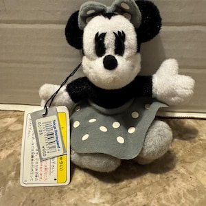 May include: A plush Minnie Mouse doll with black ears, a black and white face, and a gray bow. She wears a gray dress with white polka dots. A Tokyo Disneyland tag is attached.