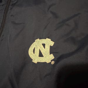 Op de afbeelding: Marineblauwe jas met een lichtgroen geborduurd "NC" logo. De jas heeft een rits en een capuchon. Het logo is een gestileerde weergave van de Universiteit van North Carolina.