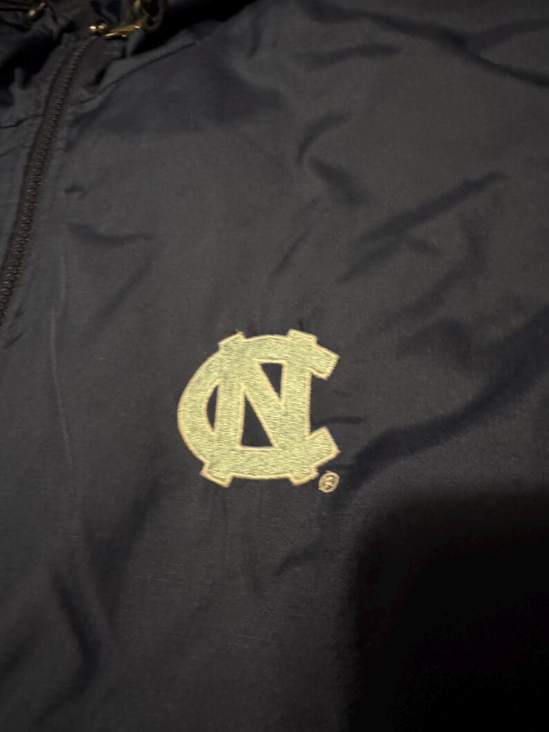 Op de afbeelding: Marineblauwe jas met een lichtgroen geborduurd "NC" logo. De jas heeft een rits en een capuchon. Het logo is een gestileerde weergave van de Universiteit van North Carolina.