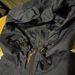 Op de afbeelding: Close-up van een marineblauwe jas met capuchon. De capuchon heeft trekkoorden met zwarte toggles en een ritssluiting. De ritssluiting heeft een goudkleurige trekker. De jas lijkt gemaakt van een licht, waterafstotend materiaal.