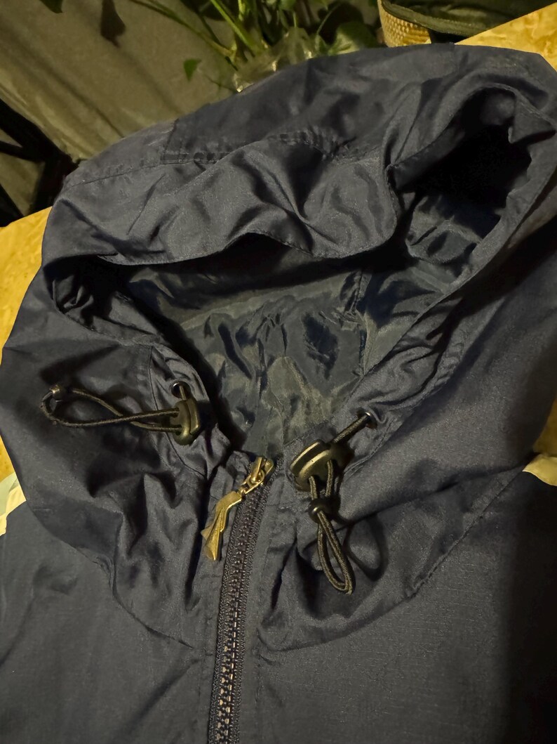 Op de afbeelding: Close-up van een marineblauwe jas met capuchon. De capuchon heeft trekkoorden met zwarte toggles en een ritssluiting. De ritssluiting heeft een goudkleurige trekker. De jas lijkt gemaakt van een licht, waterafstotend materiaal.