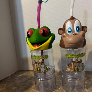 May include: Two clear plastic Rainforest Cafe cups with character lids. One lid features a green frog with red eyes and a pink straw, the other a brown monkey with blue eyes and a white straw. Both cups display the Rainforest Cafe logo and animal graphics.