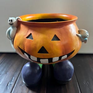 May include: A ceramic Halloween pumpkin-shaped bowl with a smiling face, triangle eyes, and white teeth. The orange bowl has two small ghost figures on the sides and rests on two purple ball-shaped feet. A fun Halloween decoration.