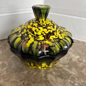 May include: A black and green glass trinket box with a yellow and brown marbled design. The lid has a knob on top.