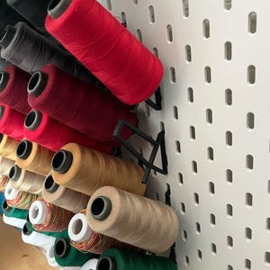 Può includere: Un pannello forato con una varietà di bobine di filo di diversi colori, tra cui rosso, nero, beige, verde e bianco. Le bobine sono organizzate in file e sono tenute in posizione da supporti in plastica nera.