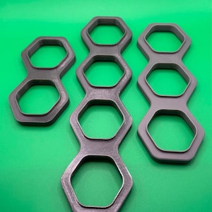 Può includere: Tre portabottiglie grigi stampati in 3D con un design a nido d'ape. I portabottiglie sono realizzati in un materiale strutturato e sono disposti su uno sfondo verde. I portabottiglie sono progettati per contenere più lattine.