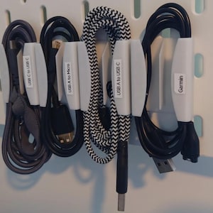 Puede incluir: Una colección de cables USB y cables de carga, cuidadosamente organizados en una pared perforada blanca. Los cables se sujetan con clips blancos, con etiquetas que indican su tipo, como "USB C to USB C" y "USB A to Micro". Cables negros, grises y estampados.