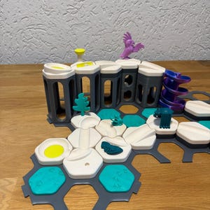 Könnte beinhalten: Ein farbenfrohes, modulares Spielzeugset mit grauen, weißen und türkisfarbenen Komponenten. Das Set enthält zylindrische Strukturen, sechseckige Kacheln und kleine dekorative Elemente wie einen lila Vogel und einen grünen Baum. Die Teile sind auf einer Holzoberfläche angeordnet.