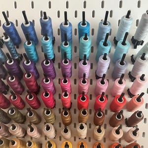 Peut inclure: Un organisateur mural avec des bobines de fil de différentes couleurs, notamment bleu, violet, rouge, marron, beige et jaune. Les bobines sont disposées en arc-en-ciel.