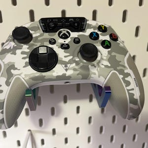 Puede incluir: Un mando Xbox blanco y gris camuflado con botones negros y un pad direccional negro. El mando tiene un diseño de camuflaje blanco y gris con un logotipo negro "RECON" en el lado derecho. El mando está montado en una pared.