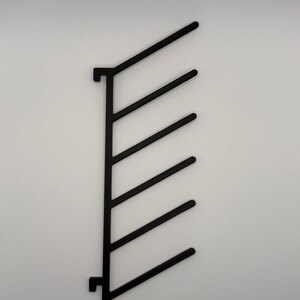 Può includere: Un espositore da parete verticale nero caratterizzato da una colonna centrale dritta con sei bracci paralleli equidistanti che si estendono diagonalmente verso destra. La struttura include due piccoli supporti a gancio in alto e in basso.