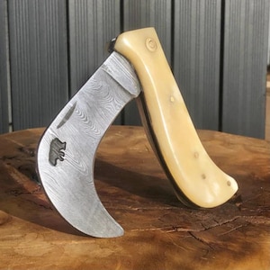 May include: A curved folding knife with a patterned blade and a cream-coloured handle. The blade features a bear emblem. The handle has a smooth, rounded shape. The knife is open, revealing its curved blade and handle design. The knife is on a wooden surface.