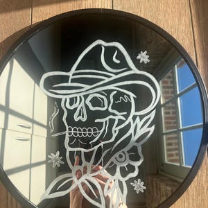 Puede incluir: Espejo redondo negro con una ilustración blanca de una calavera con sombrero de vaquero, fumando un cigarrillo, rodeada de motivos florales y estrellas. El espejo tiene un marco negro, sobre un fondo de madera.