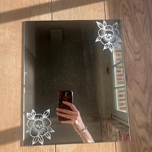 Könnte beinhalten: Quadratischer Spiegel mit geätzten weißen Blumen- und Totenkopfmotiven in den Ecken. Der Spiegel reflektiert einen Raum mit einem Fenster und einer Person, die ein Telefon hält. Er liegt auf einer Holzoberfläche.