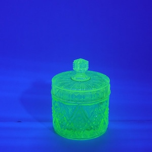 Könnte beinhalten: Ein leuchtend grünes Glasgefäß mit Deckel, vor blauem Hintergrund. Das Gefäß hat ein Rautenmuster und einen dekorativen Knauf auf dem Deckel. Das Glas scheint Vintage zu sein.
