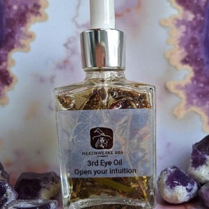 Könnte beinhalten: Eine klare Glasflasche mit 3rd Eye Oil mit einer silbernen Pipette, gefüllt mit Kräutern. Auf dem Etikett steht "Hexenwerke 888" und "Open your intuition". Die Flasche ist von lila Kristallen und einem lila-weißen Hintergrund umgeben.