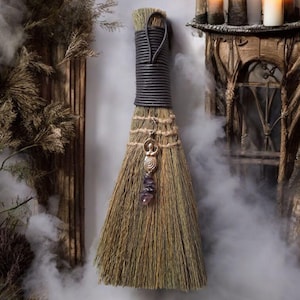 Könnte beinhalten: Ein dekorativer Besen mit einem dunkelgrauen Griff und Naturborsten. Ein silberner Anhänger mit einem Amethystkristall hängt am Besen. Der Hintergrund umfasst Kerzen und eine mystische, rauchige Atmosphäre.