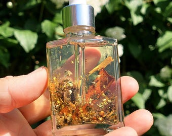 May include: A clear, square glass bottle with a silver dropper top, filled with a golden liquid and gold flakes. The bottle is held in a hand against a backdrop of green foliage and white flowers. The product appears to be a beauty oil.