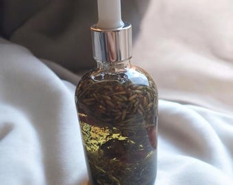 May include: A clear glass bottle filled with a golden-hued liquid and botanical elements. The bottle has a silver cap and a white dropper. The contents include green and brown herbs and red petals. The bottle sits on a white, textured surface.