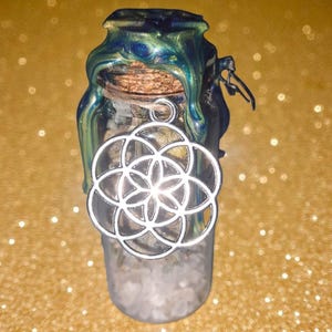 Könnte beinhalten: Kleine Glasflasche mit Korken, gefüllt mit weißen Kristallen. Die Flasche ist mit einem silbernen Anhänger mit einem geometrischen Blumendesign verziert. Die Oberseite ist mit blauem und grünem Wachs versiegelt.