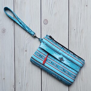 May include: A turquoise clutch with a wrist strap, featuring a Native American-inspired pattern of white feathers, red accents, and geometric shapes. The zipper pull is red, and the strap has a geometric design.