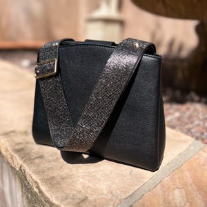 Puede incluir: Bolso bandolera negro con una correa ancha y brillante. La correa está adornada con pequeñas piedras brillantes. El bolso tiene forma rectangular con una hebilla dorada. El bolso está sobre una superficie de piedra.